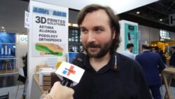 A man with a beard is interviewed, in the background several 3D printed products; copyright: beta-web | Messe Düsseldorf
