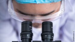 Image: Close-up of a scientist wearing protective clothing looking through a microscope; Copyright: ckstockphoto
