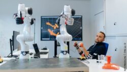 Person sitting at a table testing the controls of medical robot arms standing on the table; Copyright: TF FAU | FATHER&SUN