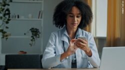 Image: A female doctor in a white coat sits at a desk and uses a smartphone; Copyright: jm_video 