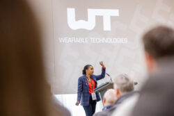 A speaker demonstrates a wearable device on stage in front of an audience at the WT | WEARABLE TECHNOLOGIES Conference 2025